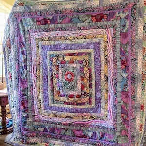 Puede incluir: Una colcha grande con un diseño cuadrado que presenta múltiples capas de tela en tonos de morado, rosa y amarillo. La colcha está hecha con una variedad de patrones y texturas, incluyendo estampados florales, remolinos y rayas.