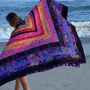 May include: A woman walks on a sandy beach holding a large, colorful quilt. The quilt is made of various shades of purple, pink, orange, and black fabric. The quilt has a geometric pattern and is decorated with floral designs.