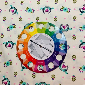 May include: A color wheel with a white background and a black arrow pointing to the color "red violet". The color wheel is surrounded by a white fabric with a repeating pattern of pink plus signs, green leaves, blue flowers, and a trophy with a blue ribbon.