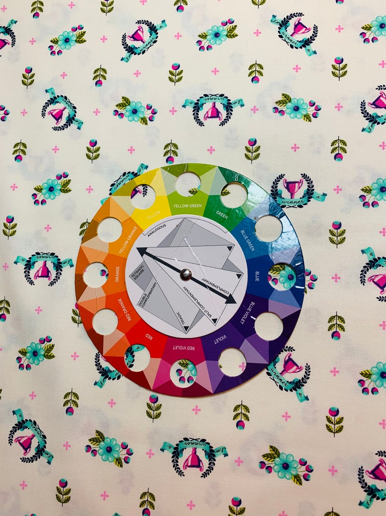 May include: A color wheel with a white background and a black arrow pointing to the color "red violet". The color wheel is surrounded by a white fabric with a repeating pattern of pink plus signs, green leaves, blue flowers, and a trophy with a blue ribbon.