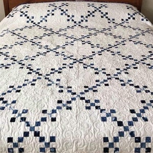 May include: A white quilt featuring a repeating lattice pattern composed of small, dark blue and white mottled squares. The fabric surface displays an intricate, swirling quilted texture throughout the white sections of the bed covering.