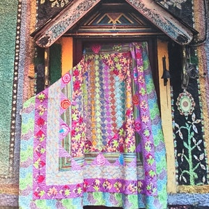 May include: A rectangular quilt featuring a vibrant patchwork design with repeating patterns of maple leaves, floral motifs, and geometric shapes in shades of purple, green, pink, and yellow. The quilt is draped over a doorway with a dark wooden frame.