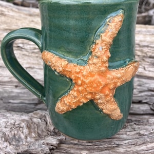 May include: A handmade ceramic mug in a deep teal glaze, featuring a raised, textured orange starfish design. The mug has a curved handle and a smooth, glossy finish, perfect for enjoying your favorite beverage. This unique mug is a great addition to any kitchen.