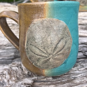 May include: A handmade ceramic mug with a sand dollar design. The mug features a two-tone color scheme, with a brown and turquoise glaze. The sand dollar is a raised, textured element on the mug's surface.