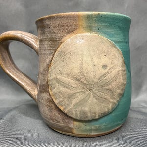 May include: Handmade ceramic mug with a sand dollar design. The mug features a brown and teal color scheme with a large, textured sand dollar affixed to the side. The handle is curved and the mug has a rustic, artisanal appearance.