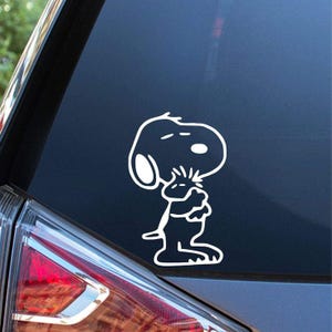 以下が含まれることがあります： 暗い背景に、自分自身を抱きしめている白いスヌーピーのデカール。デカールは、漫画のキャラクターの線画です。デカールは車のリアウィンドウにあります。