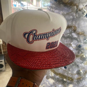 Może przedstawiać: Biała i czerwona czapka z daszkiem z napisem "Champions 2021" w niebieskich i czerwonych literach.