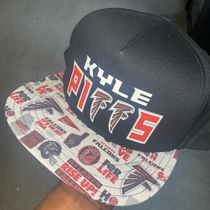 Może przedstawiać: Czarno-biała czapka bejsbolówka z czerwono-czarnym logo Atlanta Falcons i napisem "KYLE PITTS" z przodu. Czapka ma biało-szary wzór od spodu z różnymi logo i napisami Atlanta Falcons.