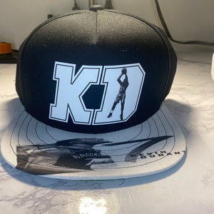 May include: Black and white baseball cap with a white "KD" logo and a silhouette of a basketball player. The cap has a white brim with a black and white graphic of a basketball player.