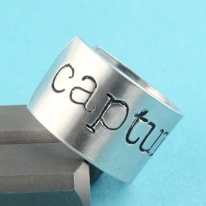 Puede incluir: Un anillo de plata con la palabra "capture" grabada.