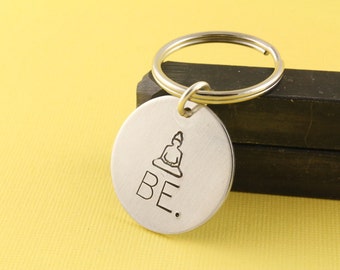 May include: Silver keychain with a round tag featuring a small engraved image of a person sitting in a meditative pose and the word "BE."