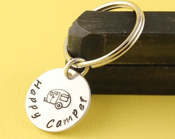 May include: Silver keychain with a round tag that says "Happy Camper" with a small camper illustration. The keychain is attached to a silver key ring.