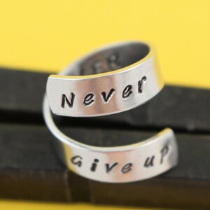 Puede incluir: Un anillo de plata con las palabras "Never Give Up" grabadas en &eacute;l. El anillo est&aacute; hecho de dos bandas que est&aacute;n retorcidas juntas.