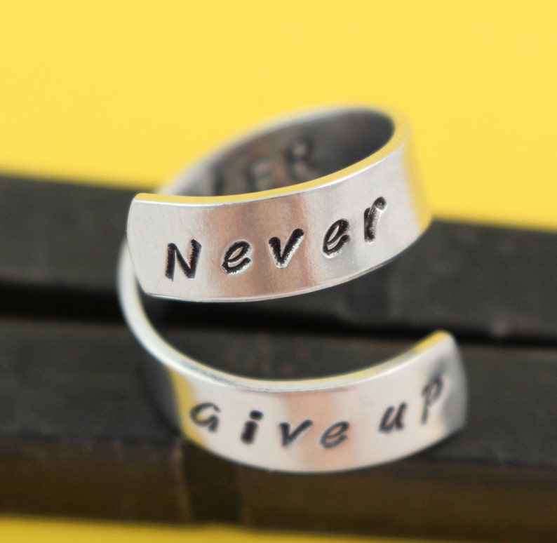 Puede incluir: Un anillo de plata con las palabras "Never Give Up" grabadas en &eacute;l. El anillo est&aacute; hecho de dos bandas que est&aacute;n retorcidas juntas.