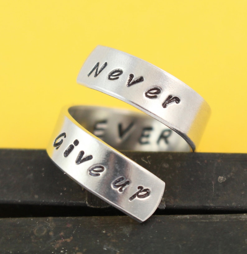 Puede incluir: Un anillo de plata con las palabras "Never give up" grabadas en &eacute;l. El anillo est&aacute; retorcido y las palabras est&aacute;n en dos bandas separadas.