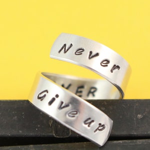 Puede incluir: Anillo de plata con las palabras "Never Give Up" grabadas. El anillo est&aacute; retorcido y las palabras est&aacute;n en dos bandas separadas.