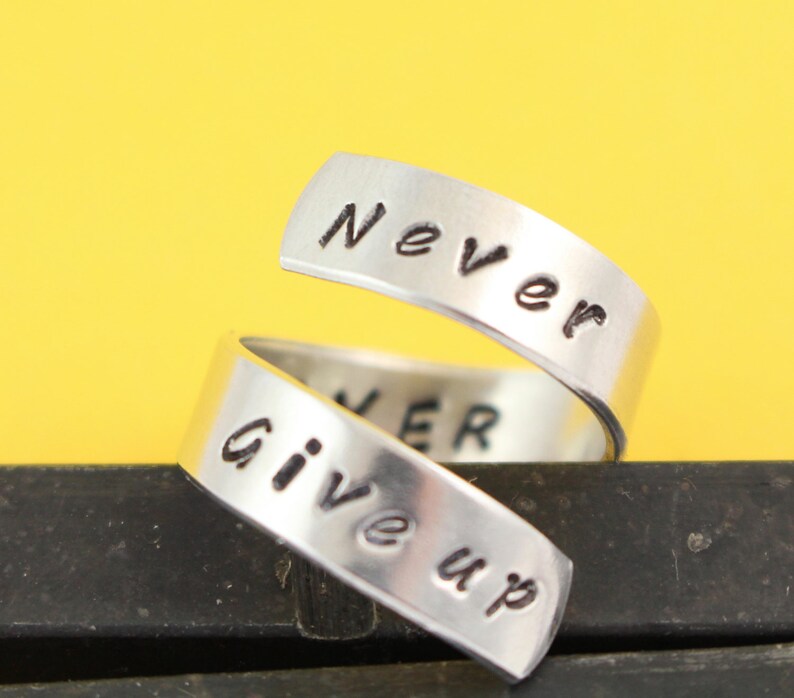 Puede incluir: Anillo de plata con las palabras "Never Give Up" grabadas. El anillo est&aacute; retorcido y las palabras est&aacute;n en dos bandas separadas.