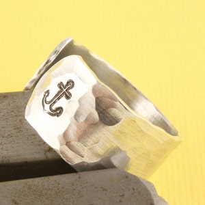 Puede incluir: Un anillo de plata con textura martillada y un diseño de ancla. El anillo es ancho y tiene una forma redondeada.