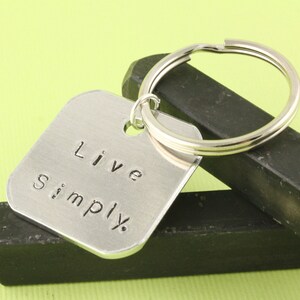 以下が含まれることがあります： 「Live Simply」と書かれた正方形のタグが付いたシルバーのキーホルダーです。
