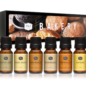 May include: A set of six 10 ml bottles of essential oil with labels for Coffee Cake, Sugar Cookie, Gingerbread, Snickerdoodle, Pumpkin Pie, and Cupcake. The bottles are arranged in a row on a white background. The box in the background says "Bakery Fragrance Collection".