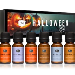 May include: A set of six 10 ml bottles of Halloween-themed essential oils. The bottles are labeled with the scents: Candy Corn, Marshmallow, Autumn Wreath, Caramel Corn, Night Air, and Pumpkin Pie. The bottles are arranged in a row on a wooden surface in front of a box with a jack-o'-lantern graphic and the text "HALLOWEEN".