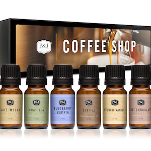 May include: A set of six 10 ml bottles of essential oil blends in a box. The bottles are labeled with the scents: Cafe Mocha, Chai Tea, Blueberry Muffin, Coffee, French Vanilla, and Hot Chocolate. The box is labeled "Coffee Shop" and features a coffee maker pouring coffee into a cup.
