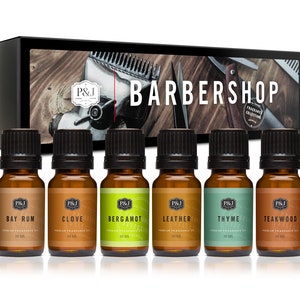 May include: A set of six 10 ml amber glass bottles with black caps, containing essential oils. The bottles are labeled with the names "Bay Rum", "Clove", "Bergamot", "Leather", "Thyme", and "Teakwood". The bottles are arranged in a row on a wooden surface. The background features a barber shop theme with a vintage barber's chair, clippers, and a razor.