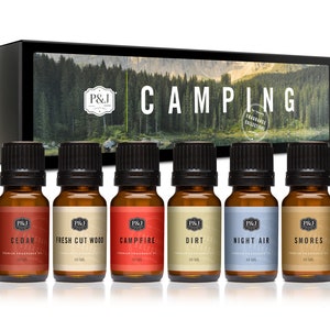 May include: A set of six 10 ml bottles of essential oil with labels that read "Cedar", "Fresh Cut Wood", "Campfire", "Dirt", "Night Air", and "Smores". The bottles are arranged in a row on a white surface. The bottles are all brown glass with black caps. The labels are all white with black text. The bottles are part of a "Camping Fragrance Collection".