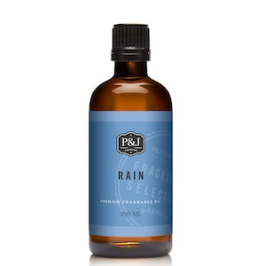 May include: A brown glass bottle with a black cap, containing a clear liquid. The label is blue with white text that reads "P&J Trading", "Fragrance Select", "Rain", "Premium Fragrance Oil", and "100 ml. Premium".