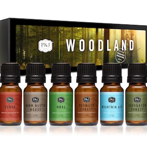 May include: A set of six 10 ml bottles of essential oil in a box. The bottles are labeled with different scents: Cedar, Warm Rustic Woods, Moss, Redwood Forest, Mountain Air, and Evergreen Forest. The box has a woodland scene on it and the text "Woodland Fragrance Collection".