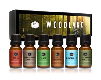 May include: A set of six 10 ml bottles of essential oil in a box. The bottles are labeled with different scents: Cedar, Warm Rustic Woods, Moss, Redwood Forest, Mountain Air, and Evergreen Forest. The box has a woodland scene on it and the text "Woodland Fragrance Collection".