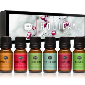 May include: A set of six 10 ml essential oil bottles in a black box. The bottles are labeled with holiday scents: Apple Cider, Candy Cane, Forest Pine, Winter Mint, Cranberry, and Mistletoe. The box has a snowy winter scene with red berries and the text "HOLIDAY FRAGRANCE COLLECTION".