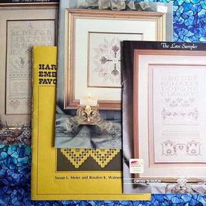 May include: Four cross stitch patterns, including "The Love Sampler" with a pink and white alphabet design, "A Grace Sampler" with a black and white alphabet design, and two other patterns with yellow and white designs. The patterns are printed on white paper and framed in black frames.
