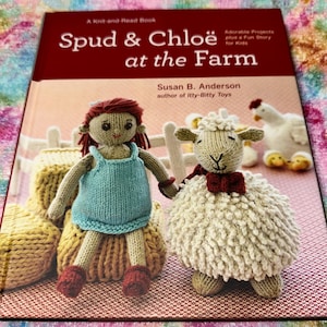May include: A softcover book titled Spud & Chloë at the Farm by Susan B. Anderson. The cover features a knitted doll in a light blue dress, a knitted sheep with a red bow, and a knitted chicken, all set against a farm-themed background.