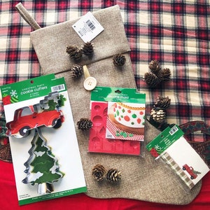 May include: A Christmas stocking, pine cones, and baking supplies are arranged on a plaid surface. The baking supplies include cookie cutters in the shape of a truck and a Christmas tree, and a silicone mold. The stocking is beige.