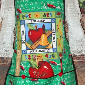 May include: A green and red apron with a school theme. It has pockets and a large apple design with the words "My Favorite" on it. The apron is covered in a pattern of letters, numbers, and school supplies.