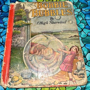 May include: Vintage children's book titled "Bobbie Bubbles" by EHugh Sherwood. The cover features an illustration of children playing with bubbles. The book has a red spine and shows signs of wear, suggesting it is an antique.
