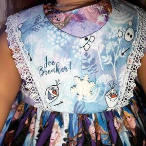 K&ouml;nnte beinhalten: Hellblaues Kleid mit wei&szlig;er Spitzenborte und V-Ausschnitt. Das Kleid zeigt einen Aufdruck von Figuren aus dem Film Frozen, darunter Olaf, Schneeflocken und den Text "Ice Breaker!"