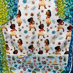 May include: White apron with a light blue pocket and a bow. The apron features a repeating pattern of a cartoon woman in a red top and grass skirt, along with other cartoon characters, flowers, and leaves.