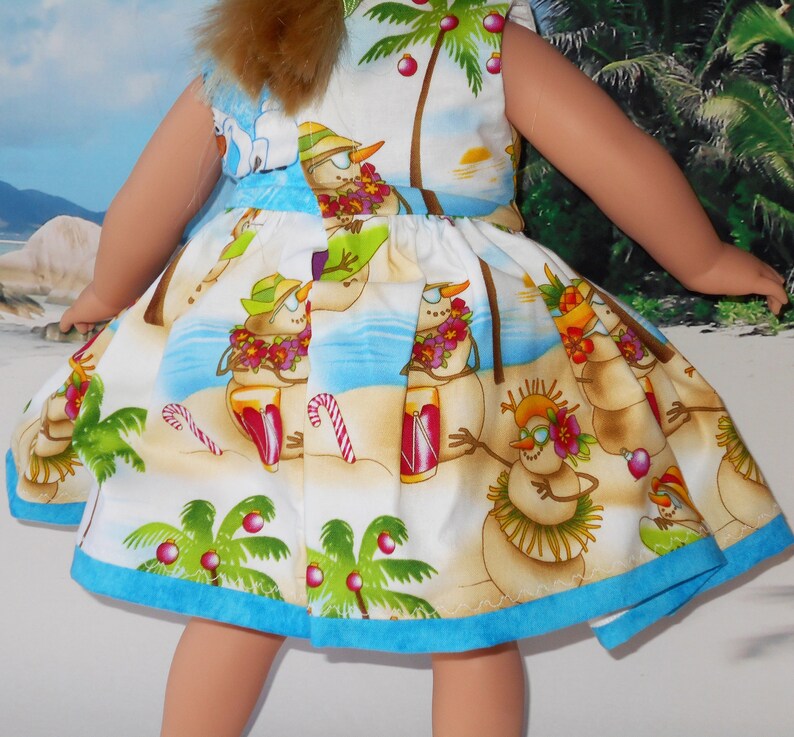 Pode incluir: Um vestido branco com uma bainha azul apresenta uma cena de praia tropical com bonecos de neve usando colares de flores e segurando bebidas. O vestido tem um tema natalino festivo com palmeiras e enfeites vermelhos.