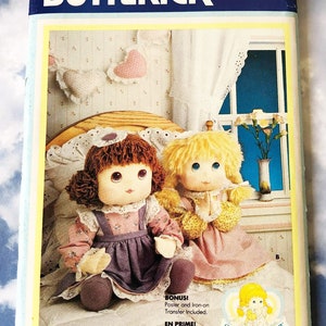 May include: A vintage sewing pattern for two dolls, one with brown hair and a purple dress, and the other with blonde hair and a pink dress. The dolls are sitting on a bed with a white lace trim. The pattern is from Butterick and is number 3049. The pattern includes a poster and iron-on transfer.