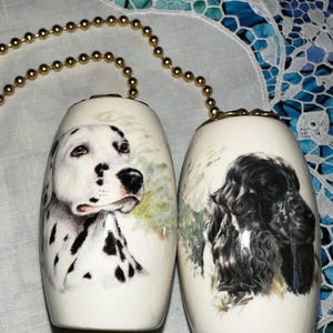 May include: A pair of white ceramic salt and pepper shakers. One shaker features a Dalmatian dog, the other a Cocker Spaniel. Both have gold-colored beaded chains attached to the top. The dogs are painted with realistic detail.