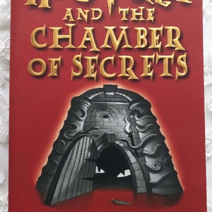 May include: A red book cover with gold lettering that reads "Harry Potter and the Chamber of Secrets" by J.K. Rowling. The cover features a black and white illustration of a stone archway with a spider inside.