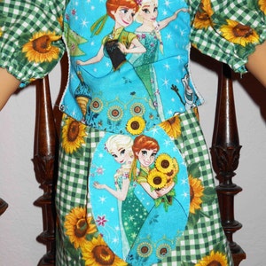 May include: A green and white gingham dress with sunflowers and a blue fabric patch featuring the characters Elsa and Anna from the movie Frozen. The fabric patch is sewn onto the front of the dress.