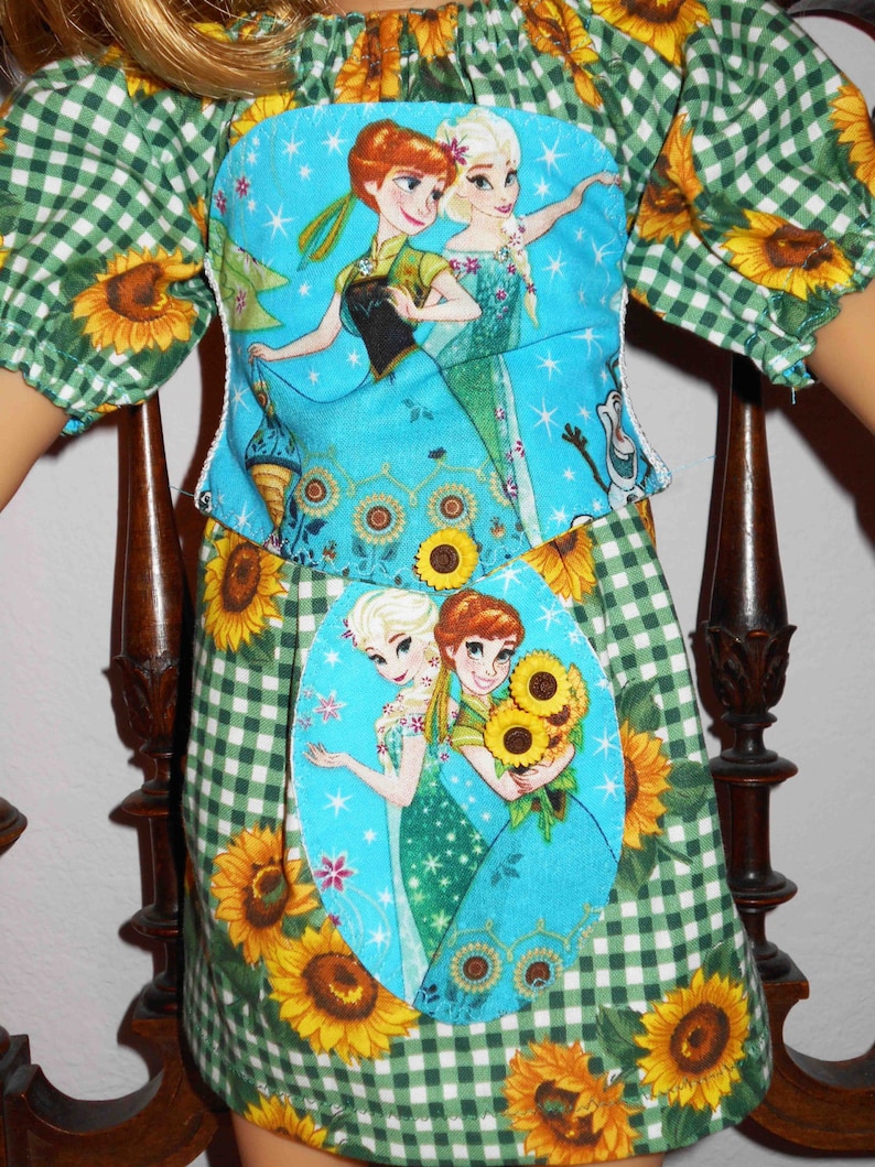 May include: A green and white gingham dress with sunflowers and a blue fabric patch featuring the characters Elsa and Anna from the movie Frozen. The fabric patch is sewn onto the front of the dress.