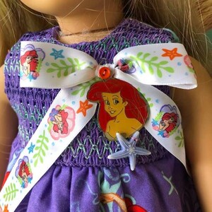 Op de afbeelding: Een paarse poppenjurk met een wit lint met Ariel-illustraties, groene bladeren en oranje zeesterren. De jurk heeft een gesmokt lijfje en een rok met Ariel-afbeeldingen. Een rode Ariel-speld en een blauwe zeester accentueren de jurk.