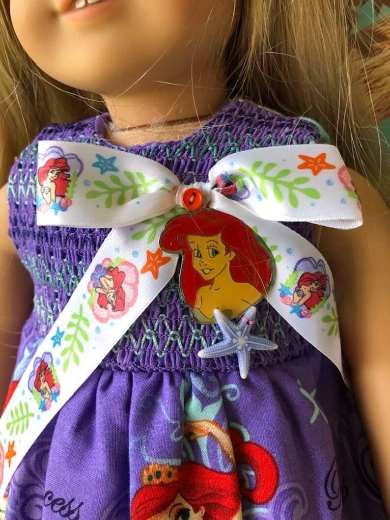 Op de afbeelding: Een paarse poppenjurk met een wit lint met Ariel-illustraties, groene bladeren en oranje zeesterren. De jurk heeft een gesmokt lijfje en een rok met Ariel-afbeeldingen. Een rode Ariel-speld en een blauwe zeester accentueren de jurk.
