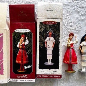 May include: Several Hallmark Keepsake Barbie ornaments from the Collector's Series are displayed. The dolls are dressed in various cultural attire, including a Russian Barbie and a Native American Barbie. The boxes are red and cream colored.