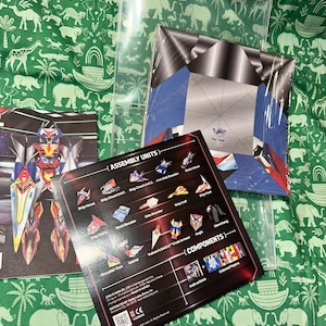May include: A paper craft model kit featuring a robot figure, including a black instruction sheet with labeled assembly units like Ship Head, Ship Chest, and Vulcan's Sword, alongside a printed sheet of colored paper components in a clear plastic sleeve.