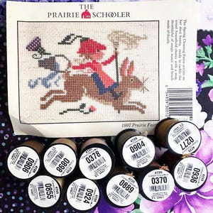 May include: A cross-stitch pattern from "The Prairie Schooler" featuring a whimsical design of a fairy riding a rabbit, with spools of embroidery floss in various colors. The pattern includes the text "1997 Prairie Fair".